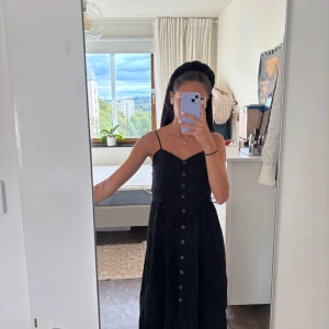 Svart långklänning med knappar - Svart långklänning med smala justerbara axelband och knappar längs hela framsidan. Klänningen har v-ringning och smockad rygg för extra komfort. Perfekt för sommardagar och enkel att styla med accessoarer.