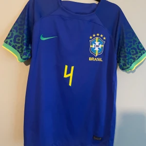 Blå Brasil Marquinhos Nike fotbollströja - Säljer en Brasilien fotbollströja med Marquinhos och nummer 4 på ryggen. Tröjan är blå med gula detaljer och har korta ärmar med leopardmönster i grönt och blått. Tillverkad i lätt Dri-Fit material som andas, perfekt för match eller träning.