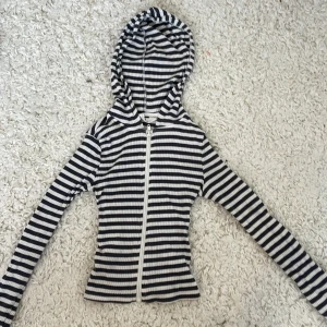 Randig hoodie med dragkedja från H&M - Säljer en svartvit randig hoodie från H&M i ribbad bomull. Tröjan har långa ärmar, huva och hel dragkedja framtill. Snygg och enkel modell som är lätt att matcha till olika outfits.