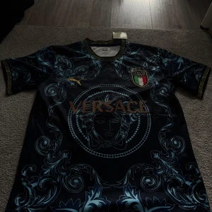 Italien x Versace x Puma fotbollströja S - Unik fotbollströja i samarbete mellan Italien, Versace och Puma. Mörkblå bas med ljusblå barockinspirerat mönster, guldiga detaljer och stor Versace-logga på bröstet. Broderat Italienskt landslagsmärke och Puma-logga. Tillverkad i lätt, ventilerande material.