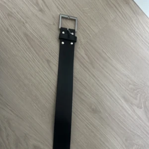 Svart skärp från ADITCHI i skinn - Snyggt svart bälte från ADITCHI i äkta skinn med en bred, stilren design och silverfärgat fyrkantigt spänne. Bältet har metallnitar vid spännet och passar perfekt till jeans eller byxor för en clean look.