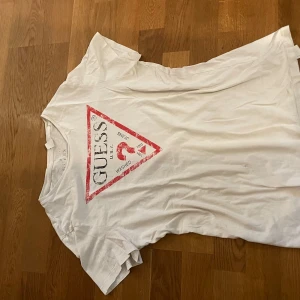 Vit t-shirt från Guess med logga - Vit t-shirt från Guess i mjuk bomull med klassisk röd triangellogga på bröstet. Modellen är kortärmad och har rund hals. Perfekt för dig som gillar stilrena och ikoniska prints. Snygg passform och lätt att matcha med jeans eller shorts.