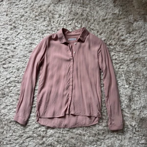 Rosa skjorta från The Shirt Factory - Säljer en stilren rosa skjorta från The Shirt Factory i storlek 36. Skjortan har klassisk krage, knappar framtill och långa ärmar med manschett. Materialet känns mjukt och lätt, perfekt för en clean och snygg look.