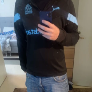 Marseille långärmad fotbollströja Puma - Säljer en svart långärmad fotbollströja från Puma med ljusblå detaljer och Marseille-logga på bröstet. Tröjan har dragkedja vid halsen och tryckt sponsor på framsidan. Perfekt för dig som vill representera OM med stil.