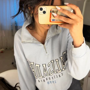 Ljusblå half zip tröja Gina Tricot - Ljusblå sweatshirt från Gina Tricot med trycket 'Palisades California 1998' framtill. Tröjan har en halv dragkedja, hög krage och långa ärmar med ribbade muddar. Oversized fit och mjukt material, perfekt för chill dagar.