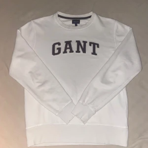 Vit sweatshirt från GANT med brodyr - Säljer en clean vit sweatshirt från GANT i storlek M. Tröjan har klassisk rund hals, långa ärmar och stor broderad GANT-logga i mörkblått framtill. Tillverkad i mjuk bomull som är skön att ha på sig. Perfekt för en chill och stilren look.