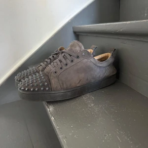 Grå mockasneakers med nitar - Snygga grå sneakers i mocka från Christian Louboutin med coola svarta nitar över tån och klassisk röd sula. Skorna har snörning och en stilren, rund tå. Perfekta för dig som vill sticka ut med en edgy detalj i din outfit.