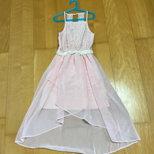 Ljusrosa långklänning med spetsdetaljer - Superfin ljusrosa långklänning med spets upptill och smala axelband. Midjan är dekorerad med blommiga detaljer och kjolen har lager på lager i tunn, flowy polyester. Perfekt för dig som vill ha en romantisk och söt look med extra volym i kjolen.