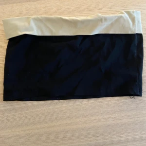 Svart och beige tubtopp - Snygg tubtopp i svart med beige kant upptill. Enkel och stilren design som passar perfekt till jeans eller kjol. Toppen är axelbandslös och har en rak bandeau-form. Perfekt för varma dagar eller lager på lager.