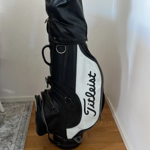 Svart och vit golfbag från Titleist - Säljer en stilren golfbag från Titleist i svart och vitt med tydlig logga på sidan. Väskan har flera fack med dragkedja och är tillverkad i skinnliknande material. Perfekt för att hålla ordning på klubborna när du är ute på banan. Bärrem ingår inte (kan köpas separat). Följande klubbor ingår: järn 4,5,6,7,8,9 och pitch. Det ingår  också trä 7 (24 grader) och trä 5 (20 grader)