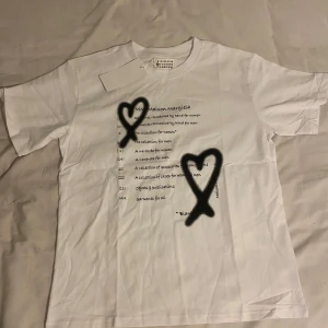 Maison Margiela vit t-shirt med tryck - Maison Margiela t-shirt i vit bomull med svart texttryck och två handmålade hjärtan på framsidan. Klassisk rund hals och korta ärmar. Snygg och clean design med coola detaljer som sticker ut.
