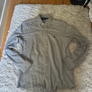Grå skjorta från Ralph Lauren - Snygg grå skjorta från Ralph Lauren i mjuk bomull. Klassisk modell med knappar hela vägen, button-down krage och broderad logga på bröstet. Perfekt för dig som gillar stilrena och tidlösa plagg med premiumkänsla.