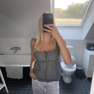 Rutig svartvit linnetopp med knytband - Supersöt linnetopp i svartvitt rutigt mönster med knytband på axlarna och liten snörning framtill. Toppen har en luftig passform och är perfekt för varma dagar. Den har en lätt volang nertill och ärmlös design som ger en chill och trendig vibe.