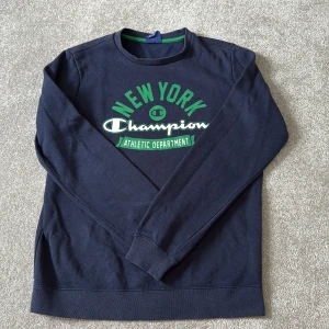 Mörkblå Champion sweatshirt L - Snygg mörkblå sweatshirt från Champion med tryck i grönt och vitt på bröstet där det står 'New York Champion Athletic Department'. Tröjan har rund hals, långa ärmar och gröna detaljer vid kragen. Perfekt för dig som gillar sportig och avslappnad stil.