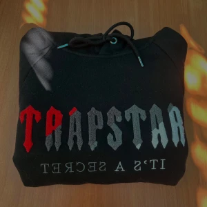 Svart Trapstar - Svart hoodie från Trapstar med stor logga i rött och grått på bröstet och texten 'IT'S A SECRET' under. Tröjan har huva med snörning och en klassisk magficka. Materialet är mjukt och känns som bomull, perfekt för chill dagar.