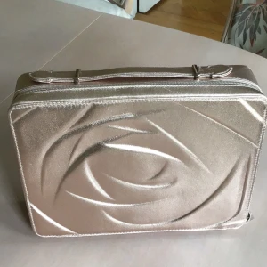 Guldig datorväska från Lancôme - Snygg och stilren smink/datorväska från Lancôme i guldigt syntetmaterial med glansig finish. Väskan har ett unikt mönster på framsidan, dragkedja runt om och ett praktiskt handtag. Perfekt för att bära din laptop, dokument eller smink med extra stil. 34x25 cm