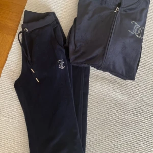 Juicy Couture set marinblå i velour - Juicy Couture set i marinblå velour med Stora glittriga strasstenar bildar loggan på ryggen och på baksidan. mindre logga framtill. Mjuk och skön, perfekt för en chill look. Klassisk streetstyle-vibe med snörning i midjan. Byxor stl XXS och hoddie stl XS