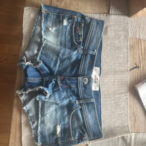 Lågmidjade jeansshorts från Hollister - Snygga lågmidjade jeansshorts från Hollister i klassisk blå denim med slitna detaljer och fransig kant. Shortsen har 4 fickor, orangea sömmar och en liten logga framtill. Perfekta för sommaren och ger en avslappnad vibe.