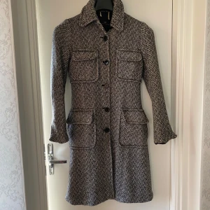 Grå ullkappa med fickor  - Snygg gråmelerad kappa från  i ullblsndning med klassiskt fiskbensmönster. Fyra stora fickor framtill, knäppning med svarta knappar och krage. Perfekt för dig som gillar en tidlös och stilren look med lite edge.