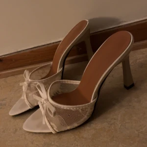 Vita spetsiga pumps med rosett - Snygga vita pumps med spetsig tå och hög klack. Skorna har öppen tå, dekorativ rosett framtill och detaljer i spets och satin. Perfekta för dig som vill sticka ut med en elegant och feminin stil.