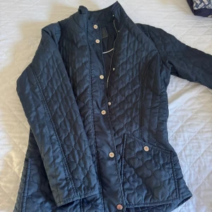 Quiltad mörkblå jacka från Cappuccini - Quiltad mörkblå jacka från Cappuccini med snygga tryckknappar och dragkedja. Jackan har lång ärm, två fickor framtill och ett stilrent mönster i tyget. Perfekt för dig som gillar klassisk men trendig stil.