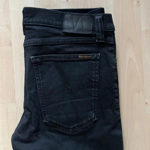 Svarta jeans från Nudie Jeans Co - Säljer ett par svarta jeans från Nudie Jeans Co i storlek 31/32. Jeansen har klassisk femficksdesign, raka ben och snygga svarta sömmar. Tillverkade i mjukt denimtyg med diskret logga på bakfickan. Perfekta för en clean och stilren look.