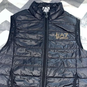 Svart dunväst från Emporio Armani EA7 - Snygg svart dunväst från Emporio Armani EA7 med guldfärgad logga på bröstet. Västen är quiltad, har hög krage och dragkedja framtill. Perfekt att slänga över en hoodie för en clean och sportig look.