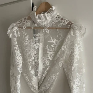Vit blommig spetsblus med volangkrage - Superfin transparent blus i vit spets med blommigt broderi och hög volangkrage. Långa ärmar med lätt puff och romantiska detaljer. Perfekt för dig som vill sticka ut med en elegant och unik look.