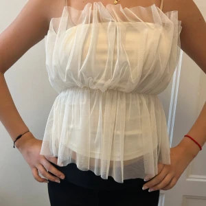 Vit tubtopp med tyll - Supersöt vit tubtopp från Vero Moda med voluminös tyll över hela toppen. Den har bandeau-modell, smala axelband och markerad midja med elastisk söm.