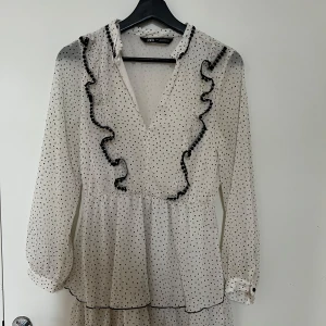 Vit prickig volangklänning från Zara - Supersöt kort volangklänning från Zara i vitt med svarta prickar. Klänningen har långa ärmar, v-ringning och fina volangdetaljer framtill. Tunt, luftigt material och markerad midja ger en flowy känsla. Perfekt för dig som gillar trenden med prickigt.