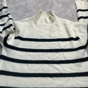 Vit och svart randig polotröja GU - Säljer en vit polotröja från GU i storlek S med svarta horisontella ränder. Tröjan har en ribbad hög krage och långa ärmar. Materialet är mjukt och stickat, perfekt för kyliga dagar. Köpt i Japan, storlek S