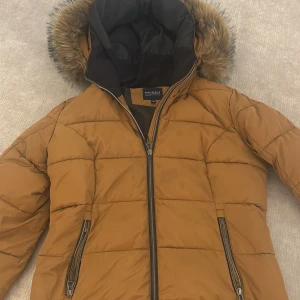 Brun pufferjacka från Race Marine - Säljer en brun pufferjacka från Race Marine med huva och fuskpäls. Jackan har två snedställda fickor en av fickorna har det gått lite sönder i, finns att se i bilden. Med dragkedja och quiltad design. Perfekt för kalla dagar och riktigt snygg till jeans eller mjukisbyxor.