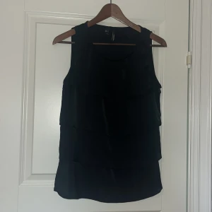 Svart ärmlös blus från Mango - Säljer en svart ärmlös blus från Mango med snygga volanglager framtill. Toppen har rund halsringning och är tillverkad i ett lätt, glansigt tyg som ger en elegant känsla. 