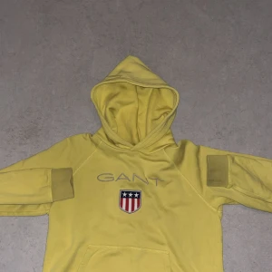 Gul hoodie från GANT med emblem - Säljer en fräsch gul hoodie från GANT med broderat emblem på bröstet och klassisk logga. Tröjan har huva, magficka och långa ärmar med ribbade muddar. Perfekt för dig som gillar sportig stil.