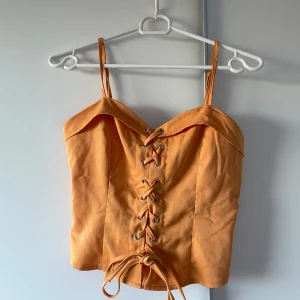 Orange korsettopp med snörning - Snygg orange korsettopp med smala axelband och cool snörning framtill. Toppen har en lätt croppad passform. Perfekt för dig som vill sticka ut med en trendig och färgstark look.