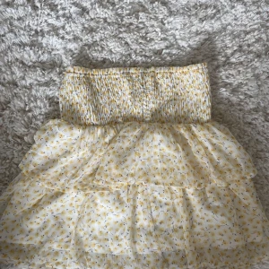 Gulblommig volangkjol med smock - Supersöt kort kjol med smock i midjan och volanger. Vit botten med små gula blommor, perfekt för sommarens alla dagar. Lätt och luftig, ger en romantisk vibe och passar perfekt till en enkel topp eller hoodie.