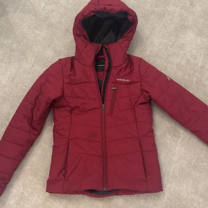 Vinröd dunjacka Peak Performance XS - Snygg vinröd vinterjacka från Peak Performance i storlek XS. Jackan har huva, dragkedja framtill och en bröstficka med dragkedja. Perfekt för kalla dagar med sin quiltade design och stilrena look. Passar dig som gillar funktion och stil i ett 