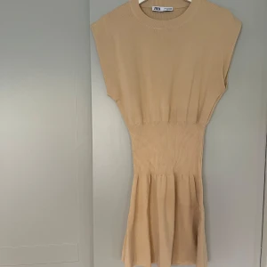 Beige stickad kortklänning från Zara - Supersnygg beige stickad klänning från Zara med rund hals och korta ärmar. Modellen har ribbad midja som ger en markerad siluett och ett lätt utställt kjolparti. Perfekt för dig som gillar enkel och stilren design med en mjuk känsla.
