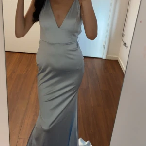 Ljusblå långklänning med snörning - Säljer en elegant ljusblå balklänning i satinliknande material med v-ringad front och smala axelband. Klänningen har öppen rygg med snygga snörningsdetaljer och en figurnära passform som ger en stilren siluett. Storlek S men passar även folk som har Xs använt engång bara och är ny