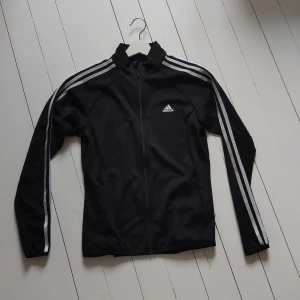 Svart Adidasjacka - Svart jacka från Adidas med klassiska vita ränder längs ärmarna och logga på bröstet. Jackan har hel dragkedja framtill, ståkrage och är tillverkad i lätt polyester. Perfekt för träning eller chill. Modell Primegreen Aeroready. Sparsamt använd.