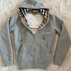 Grå Burberry hoodie med dragkedja - Säljer en grå hoodie från Burberry med klassiskt rutigt foder i huvan och insidan. Tröjan har dragkedja, snörning vid huvan och två fickor fram. Mjuk bomullskvalitet och ribbade muddar vid ärmslut och nederkant. Perfekt för chill dagar. Pris kan diskuteras vid snabb affär!!