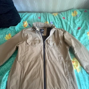Beige vindjacka från The North Face - Snygg beige vindjacka från The North Face i storlek M. Jackan har två bröstfickor med lock, hel dragkedja framtill och ventilerande meshfoder. Perfekt för dig som gillar stilren och funktionell design. Passar dig som vill ha en jacka med clean look och praktiska detaljer.