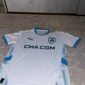 Vit Marseille fotbollströja Puma S - Snygg vit Olympique de Marseille fotbollströja från Puma med ljusblå detaljer på krage och ärmslut. Tryckt klubbmärke och Puma-logga på bröstet, samt sponsortryck. Sidorna har ett geometriskt mönster i blå och vita toner. Tillverkad i lätt polyester med DryCell-teknologi.