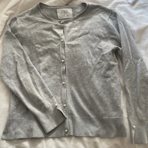 Grå kofta med pärlknappar från Zara - Snygg grå kofta från Zara. Pärlor som knappar och en på vadera arm. Använt lite. Har inget slit.