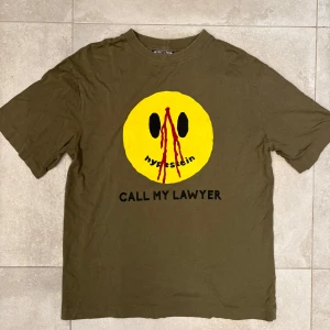 Grön t-shirt Hypestein - Olivgrön t-shirt från Hypestein med stor gul smiley och röd detalj framtill samt texten 'CALL MY LAWYER'. Relaxed oversize fit och passar M. (Nypris 500kr)