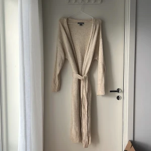 Beige lång kofta från Amisu - Säljer en beige, lång och mysig kofta från Amisu med bälte i midjan. Koftan har ribbade muddar vid ärmslut, öppen front och är stickad i ett mjukt material. Mysig och perfekt att slänga över en t-shirt när det är lite kyligt ute.