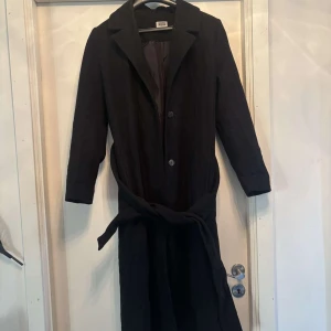 Svart kappa från Weekday med bälte - Snygg svart kappa från Weekday i storlek S. Klassisk rak modell med krage, knappar framtill och ett matchande bälte i midjan. Långärmad och fodrad för extra komfort. Perfekt för dig som gillar stilrena och minimalistiska plagg.
