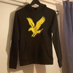 Svart lyle & scoot hoodie  - Svart hoodie med stor gul örn på bröstet. Tröjan har huva, lång ärm och en klassisk magficka framtill. Materialet känns som mjuk bomull eller polyester, perfekt för chill dagar. Snygg kontrast mellan svart och gult som sticker ut.