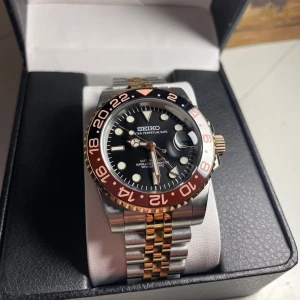 Seiko GMT-Master 2 ”Rootbeer” Mod - Helt nybyggd Seiko GMT-Master 2 ”Rootbeer” Mod  Specifikationer: Storlek: 40mm Urverk: Nh34 (Automatiskt med GMT Funktion) Material: 904L Rostfritt Stål (Two-Tone Rosé/Silver) Glas: Sapphire Glas med Förstoring vid Datum Boett: Rootbeer färger på Bezel Tavla: Svart/Rosé detaljer med Datum Armband: Viklås och Justerbart (Jubilee med Two-Tone) Krona: Screw Down Vattentålighet: 30 Meter  En klassiker Rootbeer! Med denna klockan så kommer du att få många fina kommentarer! DM vid Intresse.