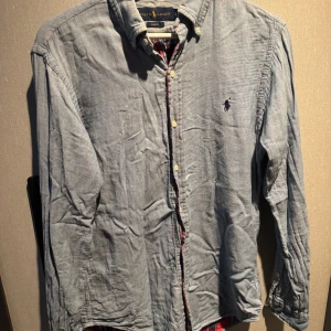 Ljusblå skjorta från Ralph Lauren - Snygg ljusblå skjorta från Ralph Lauren i slim fit-modell. Skjortan har klassisk krage, knappar framtill och det ikoniska broderade logot på bröstet. Tillverkad i mjuk bomull och har långa ärmar. Perfekt för dig som gillar stilrena och tidlösa plagg.